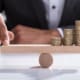 Close-up Of A Businessperson's Hand Balancing Stacked Coins On Wooden Seesaw With Finger Over Desk