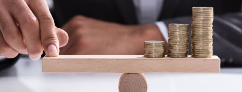 Close-up Of A Businessperson's Hand Balancing Stacked Coins On Wooden Seesaw With Finger Over Desk